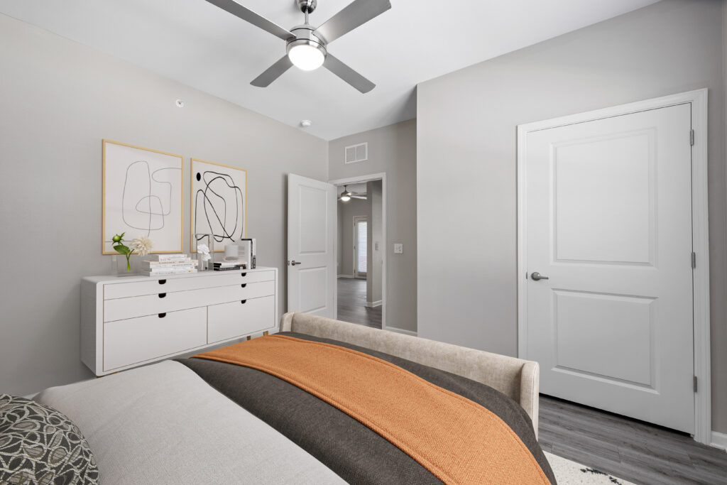 A beige bedroom at Le Jolliet in Lake Charles, LA, featuring a comfortable bed and a white dresser topped with two elegant paintings.